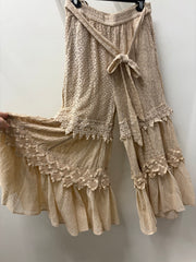 A pair of beige wide-leg bohemian pants with intricate lace and crochet patterns, tiered ruffles, and a tie-front waist, shown hanging against a plain wall. Desert Dream Cloud Pants by Young Threads Robin Boutique